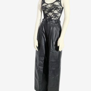 May include: A mannequin displays a black lace vest top and high-waisted black leather trousers. The vest top features a scoop neckline and a floral lace design. The trousers are wide-legged with a pleated front.