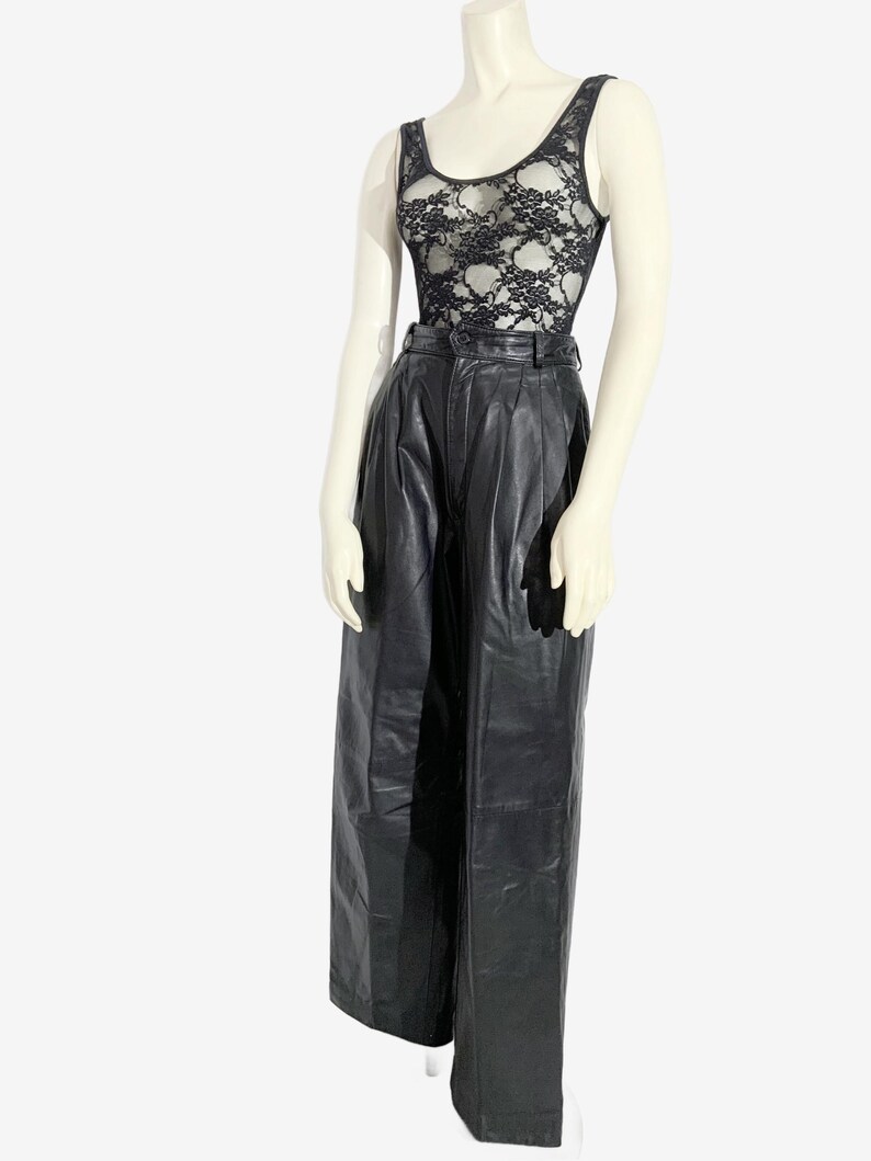 May include: A mannequin displays a black lace vest top and high-waisted black leather trousers. The vest top features a scoop neckline and a floral lace design. The trousers are wide-legged with a pleated front.