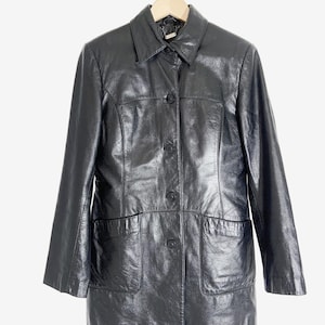 May include: Black leather jacket with a collared neckline, button-up front, and two front pockets. The jacket is hanging on a wooden hanger against a white background. The jacket has a shiny finish.