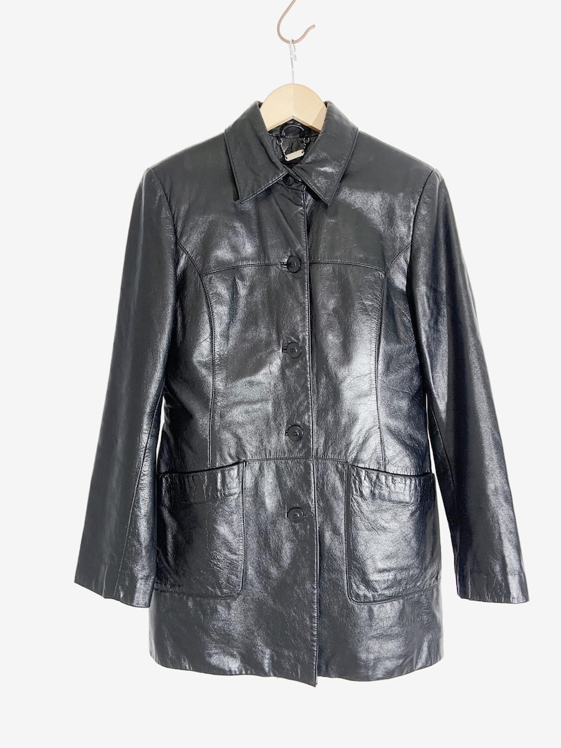 May include: Black leather jacket with a collared neckline, button-up front, and two front pockets. The jacket is hanging on a wooden hanger against a white background. The jacket has a shiny finish.