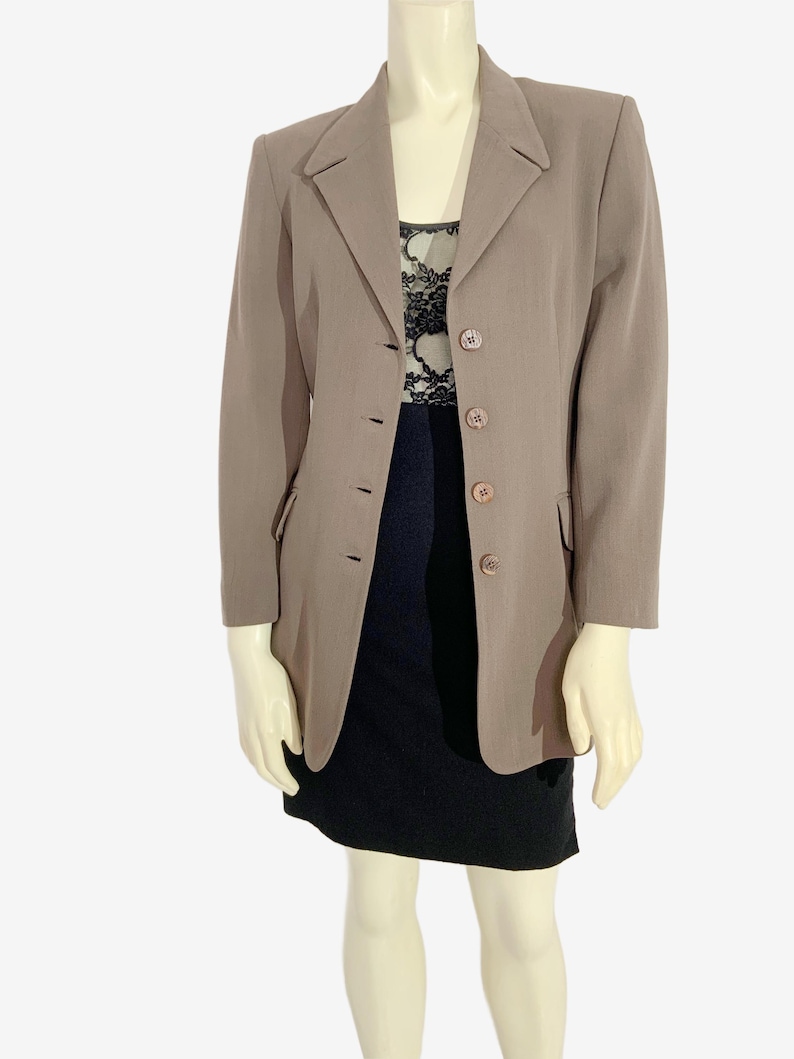 May include: A taupe blazer with a notched lapel and three-button closure. The blazer is worn over a black dress with a lace top. The mannequin is wearing a black skirt. The blazer has two front pockets.