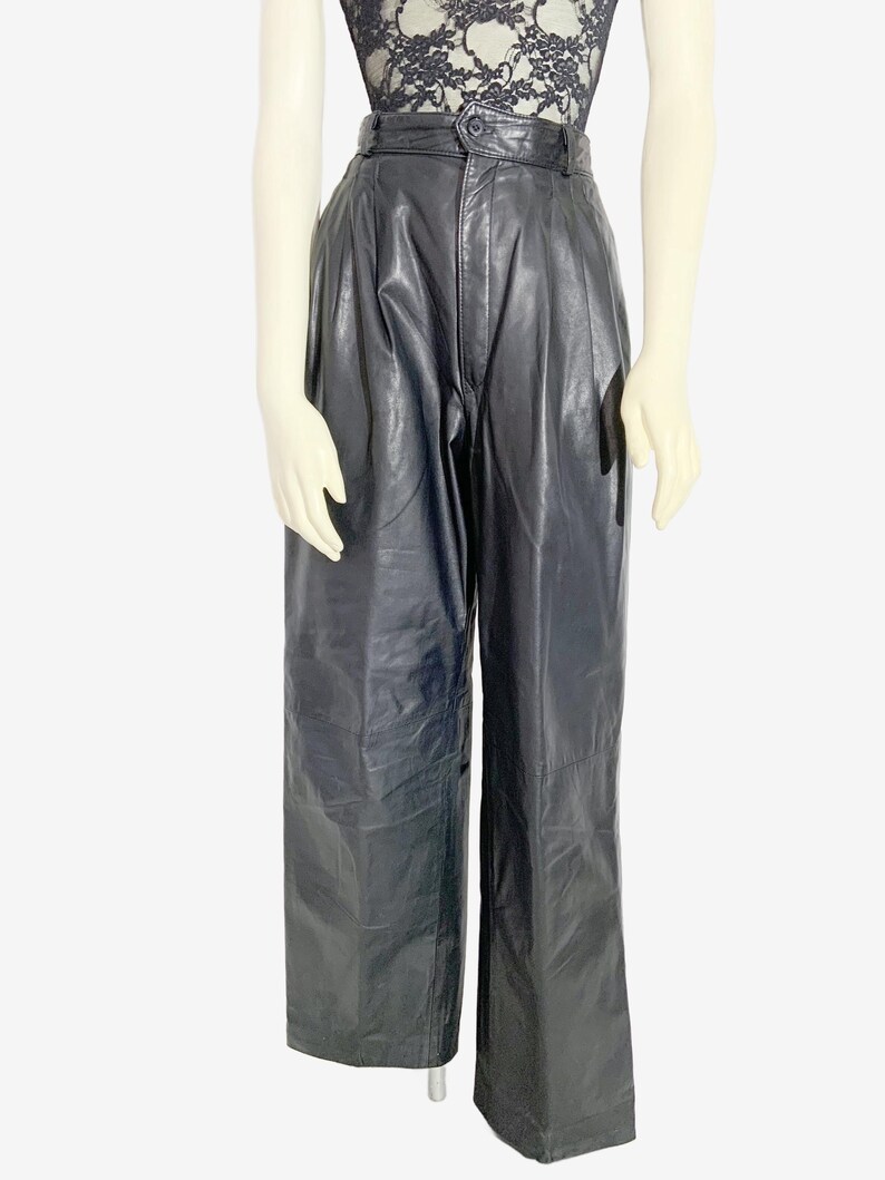 May include: Black leather trousers with a high waist and wide legs. The trousers have a button closure and pleats at the waist. The model is wearing a black lace top.