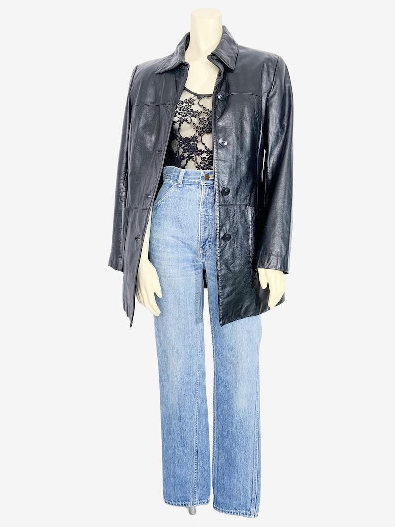 May include: A black leather jacket with a button-up front, worn over a black lace top and light blue denim jeans. The jacket has a collared neckline and long sleeves. The jeans are high-waisted with a wide leg.