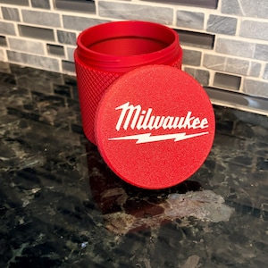 Peut inclure: Un récipient cylindrique rouge vif avec un extérieur texturé à motif de losanges et un couvercle circulaire assorti. Le couvercle présente le logo blanc Milwaukee avec un graphique en forme d'éclair. Le récipient est ouvert et creux.