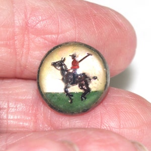 May include: A small, round button with a painted equestrian scene. The image features a rider in a red coat on a black horse, set against a green and yellow background. The button has a metallic, possibly brass, rim.
