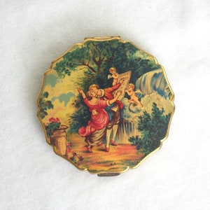 May include: A vintage compact mirror with a scalloped edge and a gold-tone frame. The lid features a painted scene of figures in a garden with a waterfall. The colors include red, green, yellow, and gold.