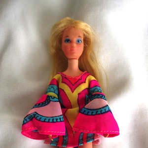 May include: A vintage doll with blonde hair and blue eyes, wearing a colourful dress. The dress features a pink, yellow, and blue pattern with flared sleeves. The doll is standing on a white surface.