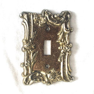 Puede incluir: Cubierta de interruptor de luz ornamentada, de estilo vintage, con una sola abertura para palanca. La cubierta presenta un centro de color bronce y un marco decorativo de color dorado con detalles florales y de volutas. La cubierta es rectangular.