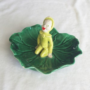 Può includere: Figurina vintage in ceramica di un elfo seduto in abito giallo con un sorriso rosso, appoggiato su un piatto verde a forma di foglia. La figurina ha un viso bianco ed è un oggetto decorativo stravagante.