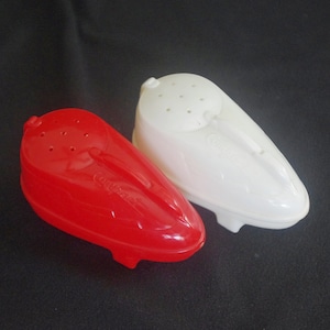 May include: Two plastic salt and pepper shakers. One is bright red, the other is white. Both have a rounded, streamlined shape with a series of holes on top for dispensing. The shakers are on a black surface.