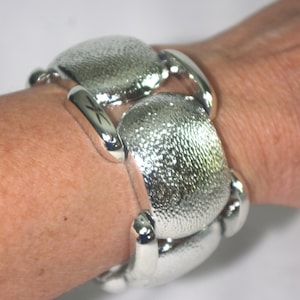 Puede incluir: Un brazalete plateado con un diseño ovalado texturizado. El brazalete está compuesto por múltiples eslabones conectados, cada uno con una superficie elevada y con hoyuelos. Los eslabones están conectados por barras plateadas curvas y pulidas. El brazalete se lleva en la muñeca.