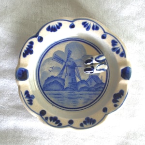 Può includere: Un posacenere rotondo in ceramica con bordo smerlato, caratterizzato da una scena dipinta a mano in blu e bianco di un mulino a vento vicino all'acqua. Il bordo è decorato con motivi floreali blu e all'interno poggia un piccolo paio di zoccoli in ceramica.