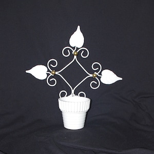 May include: White metal wall planter with a decorative leaf and scroll design. The planter has a small, ribbed pot for holding plants. The metalwork features a diamond shape with leaf accents and small gold-colored spheres.