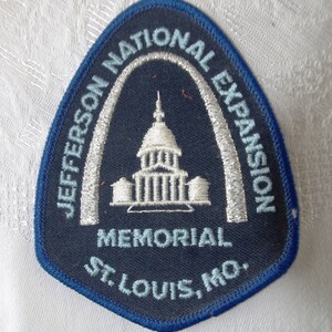 May include: A navy blue patch with a white and light blue embroidered design. The patch features the text "JEFFERSON NATIONAL EXPANSION MEMORIAL ST. LOUIS, MO." and an image of a building under an arch.