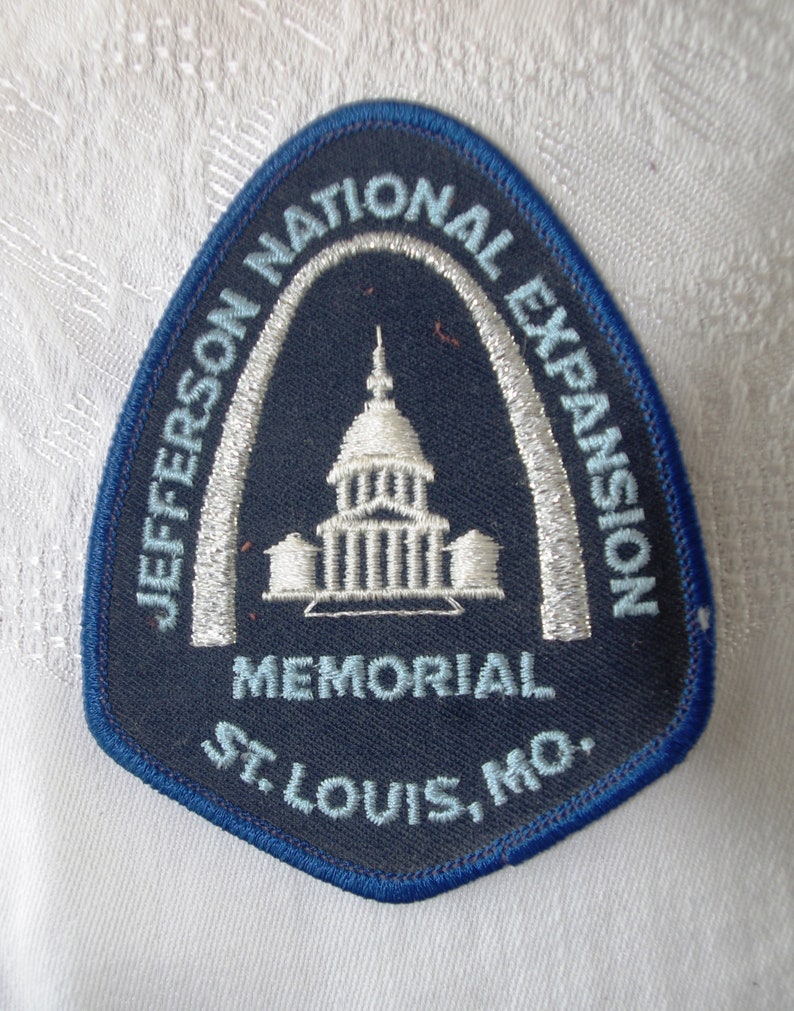 May include: A navy blue patch with a white and light blue embroidered design. The patch features the text "JEFFERSON NATIONAL EXPANSION MEMORIAL ST. LOUIS, MO." and an image of a building under an arch.