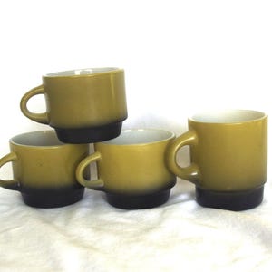 Puede incluir: Un conjunto de cuatro tazas de cerámica con un diseño degradado que transita desde una base marrón oscuro hasta una parte superior de color verde oliva apagado. Cada taza tiene un asa redondeada y lisa y un interior blanco, dispuestas en grupo.