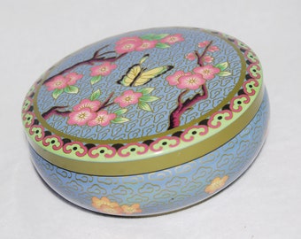 May include: Oval-shaped decorative box with a light blue background and a floral design. The lid features pink flowers, green leaves, and a yellow butterfly. The box has a gold and green trim.