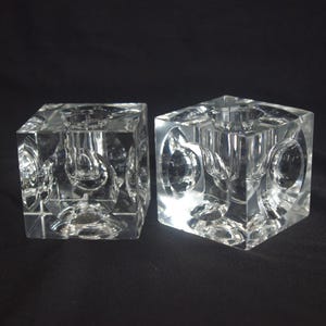 May include: Two clear, square glass candle holders. Each cube-shaped holder has a circular indentation on each side and a central opening for a candle. The candle holders are displayed against a black background.