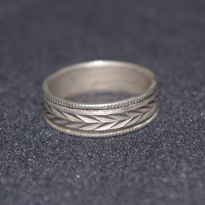 Könnte beinhalten: Silberring mit detailliertem Blattmuster um das Band. Der Ring hat einen strukturierten Rand an den oberen und unteren Kanten. Der Ring ist ein einfacher Bandstil.