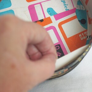 May include: A vintage, round tin with a colorful interior featuring a retro geometric pattern in pink, orange, and blue. The tin's edge is black with a decorative border. A hand is peeling back a section of the tin's interior.