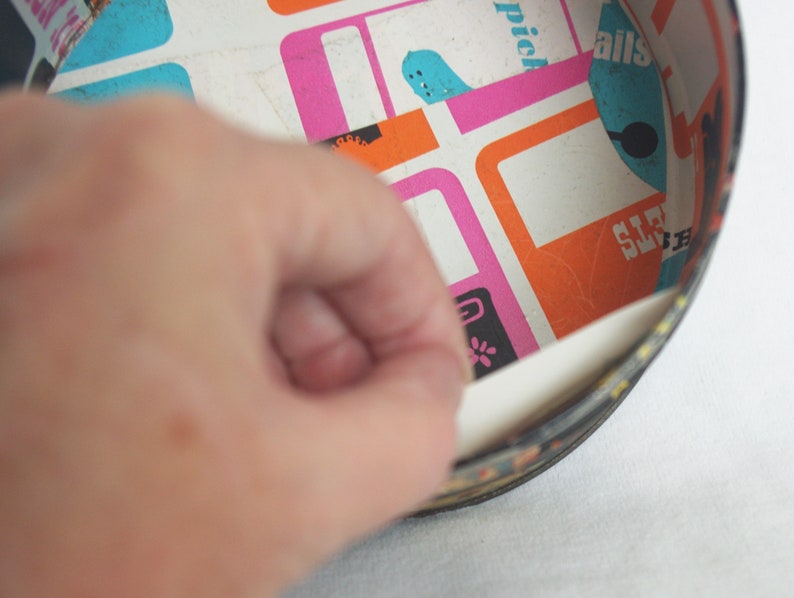 May include: A vintage, round tin with a colorful interior featuring a retro geometric pattern in pink, orange, and blue. The tin's edge is black with a decorative border. A hand is peeling back a section of the tin's interior.