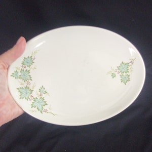 Könnte beinhalten: Ovale Servierplatte in Creme mit einem Blatt- und Beerenmuster in Grün und Rot. Das Design befindet sich auf der linken und rechten Seite der Platte. Die Platte wird in einer Hand gehalten.