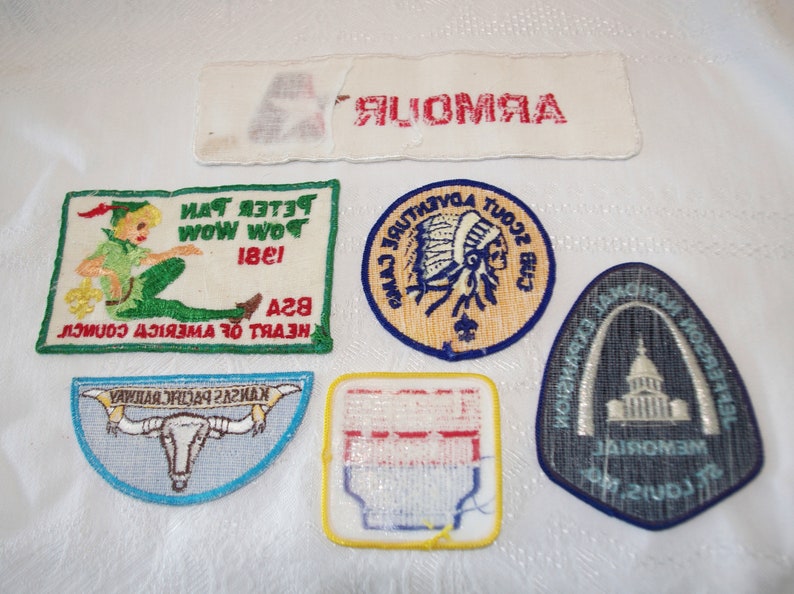 May include: A collection of vintage embroidered patches, including a Peter Pan design, a Native American scout emblem, and a Kansas Pacific Railway patch. The patches are in various shapes and colors, with text and detailed imagery.