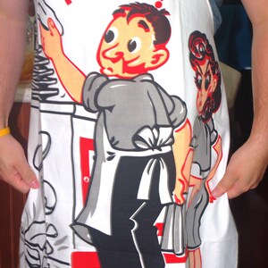 May include: White apron with a cartoon chef illustration and the text "King of the Kitchen!" in red. The apron features a retro design with a chef holding a spatula and a woman in a gray dress. The apron is a kitchen accessory.