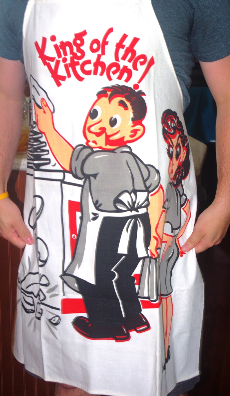 May include: White apron with a cartoon chef illustration and the text "King of the Kitchen!" in red. The apron features a retro design with a chef holding a spatula and a woman in a gray dress. The apron is a kitchen accessory.