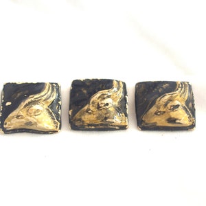Può includere: Tre piastrelle quadrate in ceramica in stile vintage. Ogni piastrella presenta una testa di ariete color oro su sfondo nero. Le piastrelle sono disposte in fila su uno sfondo bianco. La testa di ariete è dettagliata con corna e lineamenti.