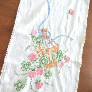 May include: White embroidered table runner with a floral design. The embroidery features pink flowers, green clovers, and a brown basket-like structure, accented with a blue ribbon. The edges are finished with a white crocheted trim.