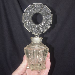Pu&ograve; includere: Un flacone di profumo in vetro trasparente con un tappo decorativo a forma di stella. La bottiglia ha una base quadrata e un corpo sfaccettato. Il tappo &egrave; un design circolare con un'apertura centrale. La bottiglia &egrave; tenuta in mano su sfondo nero.