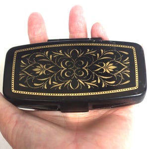 Könnte beinhalten: Ein schwarzer rechteckiger Kompakt mit einem goldenen Blumendesign. Der Kompakt wird in einer Hand gehalten und hat einen goldenen Rand. Das florale Design ist symmetrisch und detailliert, mit einem Punktmuster.