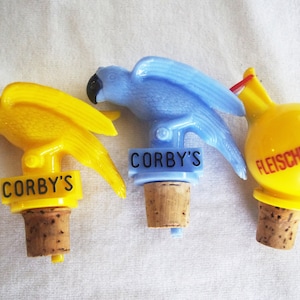 May include: Three vintage bottle stoppers. Two are parrot-shaped, one yellow and one blue, with "CORBY'S" printed on them. The third is a yellow sphere with "FLEISCHMANN'S" printed on it. All have cork bases.