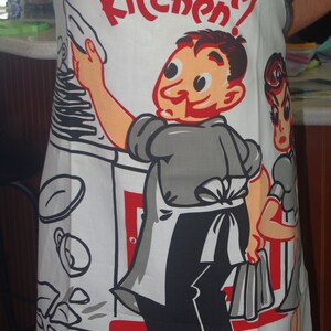 May include: White apron with a retro cartoon illustration. The apron features a chef character with the text "King of the Kitchen!" in red. The design includes a woman and kitchen utensils.