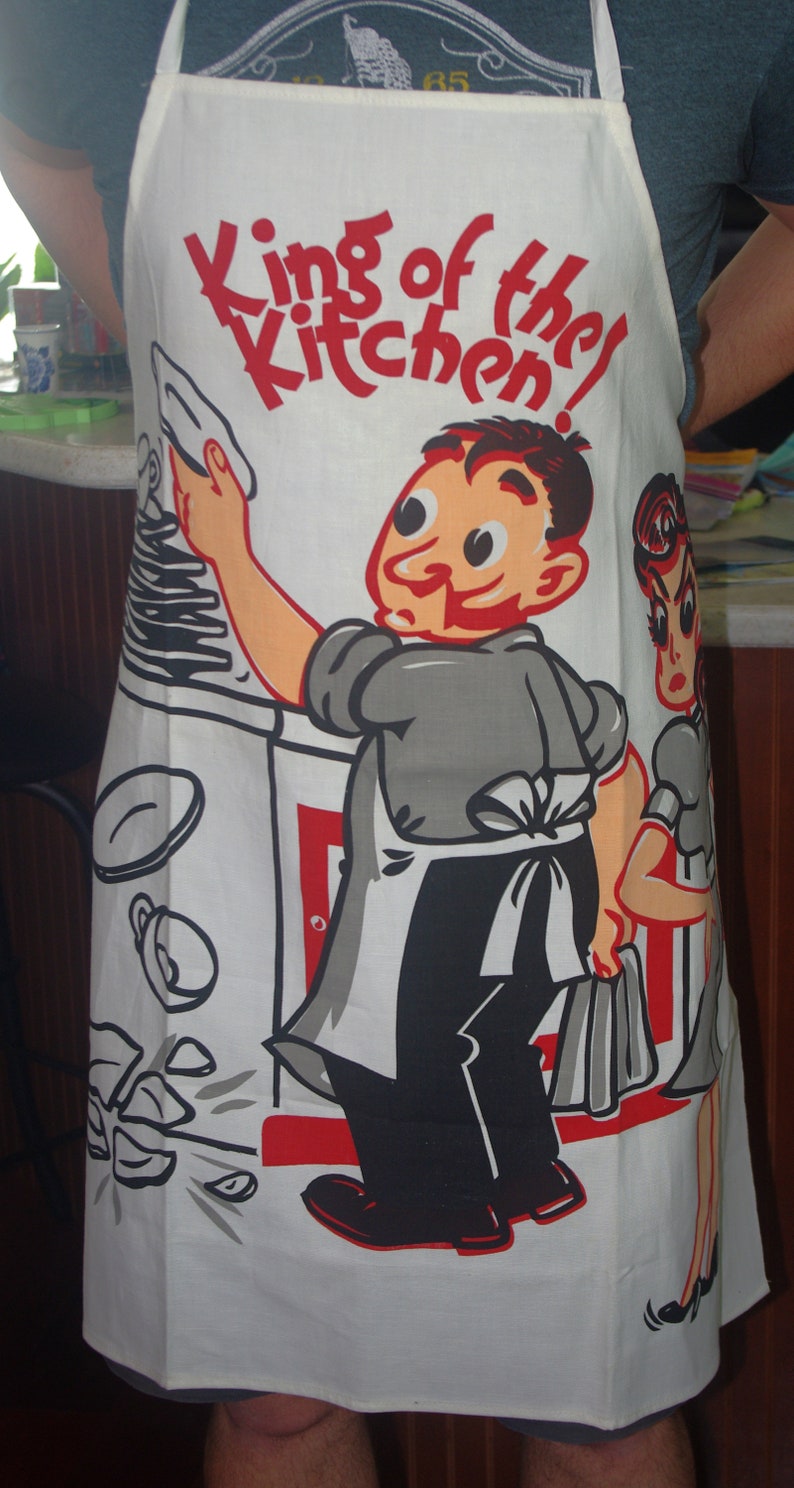 May include: White apron with a retro cartoon illustration. The apron features a chef character with the text "King of the Kitchen!" in red. The design includes a woman and kitchen utensils.