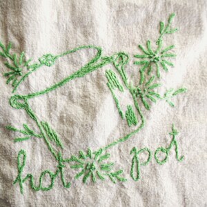 Puede incluir: Diseño bordado en verde de una olla caliente con detalles florales sobre tela blanca. Las palabras "hot pot" están bordadas debajo. El bordado es de estilo sencillo y artesanal.