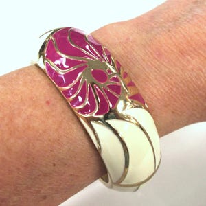 May include: A wide, gold-toned bangle bracelet with a floral design. The bracelet features a magenta and gold floral pattern on the top, with cream-colored enamel and gold accents. The bracelet is approximately 2.5 inches wide.