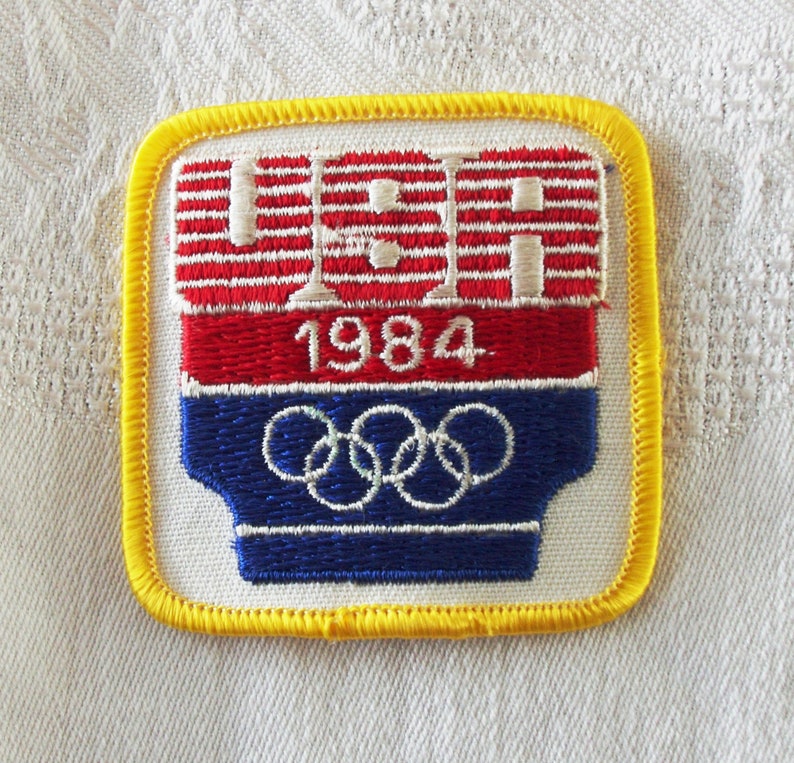 May include: A vintage patch featuring the text "USA" in white, red and blue stripes above the year "1984" and the Olympic rings. The patch has a yellow border and is likely a souvenir or collectible item.