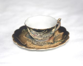 May include: A vintage teacup and saucer set. The cup and saucer are black with gold trim and ornate dragon designs. The cup has a white interior and a small handle. The saucer has a scalloped edge.