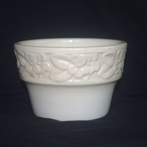 May include: A white ceramic planter with a floral embossed design around the rim. The planter has a cylindrical shape with a slightly flared base. The background is a solid black.