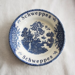 May include: A white and blue decorative plate with the word "Schweppes" in a curved font at the top and bottom. The plate features a blue and white Asian-inspired landscape design with a pagoda, bridge, and trees.