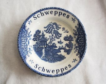 May include: A white and blue decorative plate with the word "Schweppes" in a curved font at the top and bottom. The plate features a blue and white Asian-inspired landscape design with a pagoda, bridge, and trees.