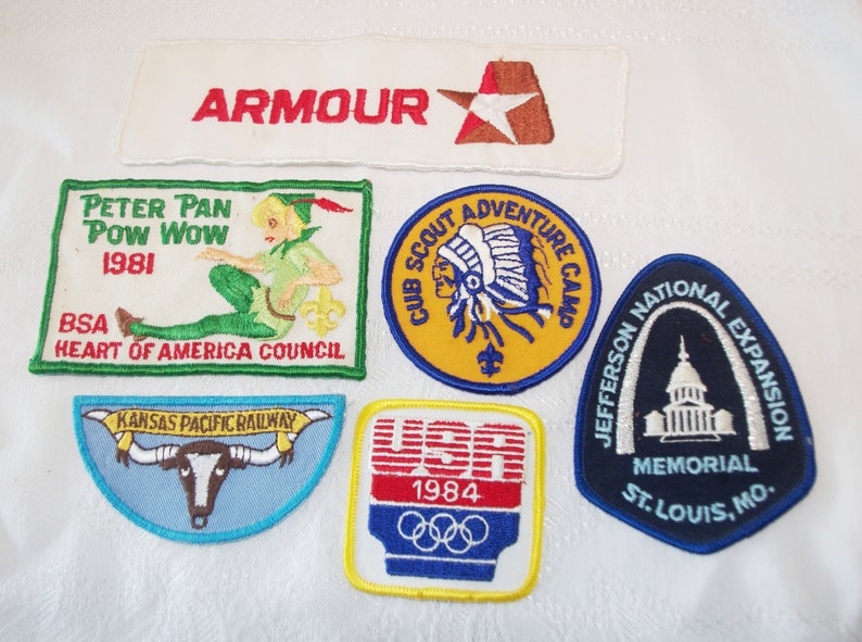 May include: A collection of vintage embroidered patches. Includes a white patch with "ARMOUR" in red, a Peter Pan patch, a Cub Scout Adventure Camp patch, a Kansas Pacific Railway patch, a USA 1984 Olympics patch, and a Jefferson National Expansion Memorial patch.