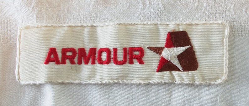 May include: Cream-colored embroidered patch with the word "ARMOUR" in red letters. A brown and red star design with a white star is on the right side of the patch. The patch has a white border.
