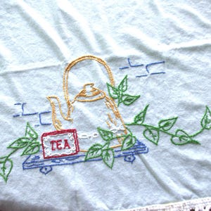 May include: Embroidered tea towel with a teapot design. The towel features a gold teapot, a red and white "TEA" box, and green leaf vines. Blue brick-like shapes are also embroidered on the light blue fabric. The bottom edge has a white lace trim.