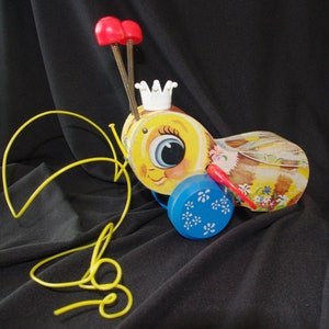 May include: A vintage pull-along toy bee with a yellow body, smiling face, white crown, and red antennae. It has a blue drum with white flower designs and a yellow pull string. The toy is designed to be pulled along.