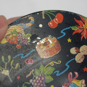 May include: A black, round decorative tin with a festive design. The tin features illustrations of cherries, grapes, a cake, nuts, a pineapple, and a poinsettia. Blue ribbons and gold stars accent the design.