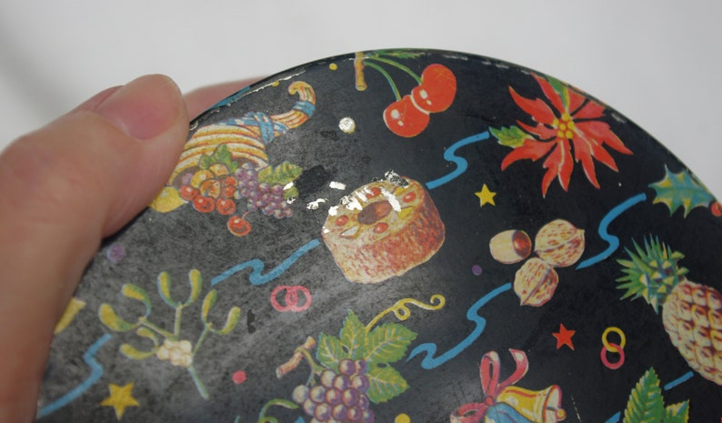 May include: A black, round decorative tin with a festive design. The tin features illustrations of cherries, grapes, a cake, nuts, a pineapple, and a poinsettia. Blue ribbons and gold stars accent the design.
