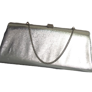 Può includere: Pochette argentata con tracolla a catena. La borsa rettangolare ha una superficie strutturata e una chiusura a scatto. La catena è di colore argento scuro. La borsa è su sfondo bianco.